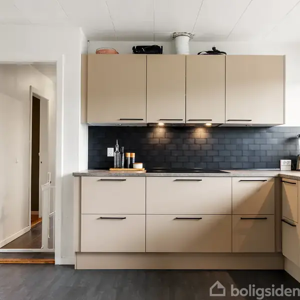 Køkken med beige skabe og skuffer, sort bordplade og backsplash. Under skabene er der tre spots tændt. Ved siden af er en åben dør, og vindue med planter.