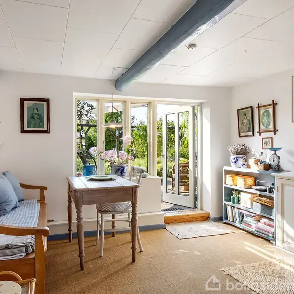 En hyggelig stue med en træbænk og et bord foran store vinduer, der åbner ud til en grøn have. Væggene har diverse billeder og dekorationer.