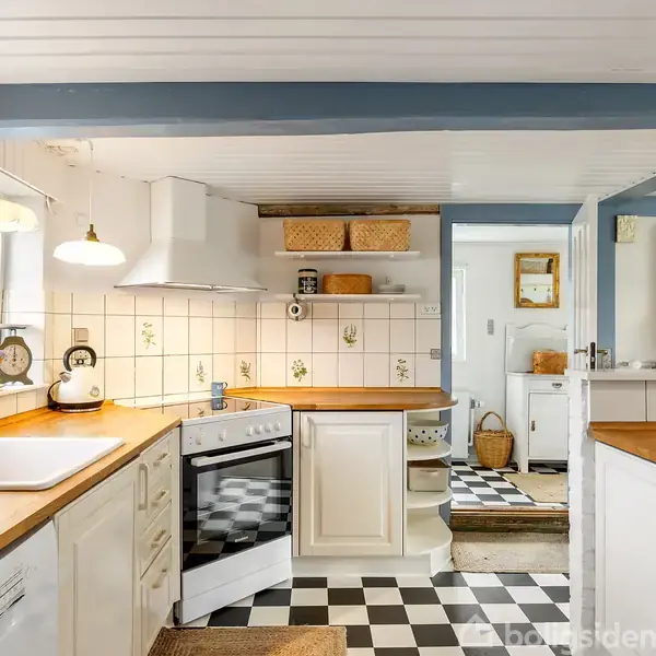 Køkken med hvide skabe og træbordplader, hvor en elkedel står ved kogepladen. Flisebelagte vægge med blomstermotiver og sort-hvidt ternet gulv. Naturligt lys fra vinduerne skaber en lys atmosfære.