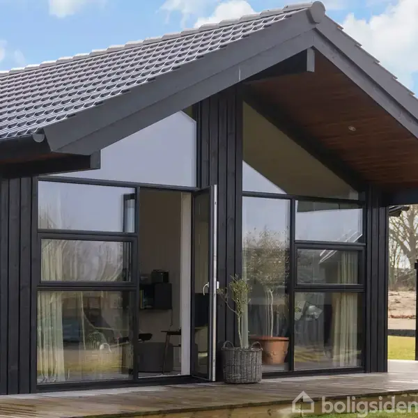 Et moderne sommerhus med store glaspartier og sort træbeklædning. Det står på en terrasse med planter i keramikpotter. Omgivelserne er en grøn have med træer i baggrunden.