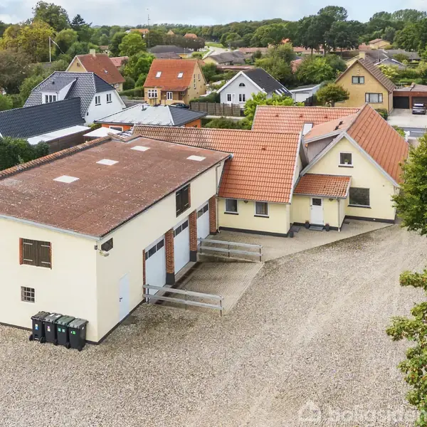 Et stort hvidt hus med rødt tegltag ligger i et villakvarter, omgivet af andre huse og grønne træer. Grusbelagt gårdsplads foran bygningen.