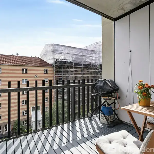 Balkon med sorte gelændere, fyldt med møbler og planter, tilknyttet en moderne lejlighedsbygning. Over- og underliggende balkoner tilføjer dybde til den travle byscene.