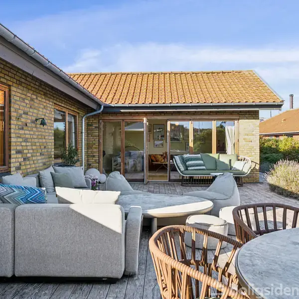 Stue i udendørs miljø: Flotte, lysegrå sofaer og et rundt bord står på en træterrasse i solskin. Baggrunden viser et murstenshus med store vinduer og grønne buske.