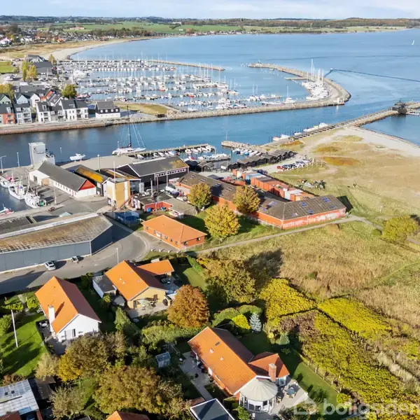 Kystby med både i havn, rødtaget huse i forgrunden. Bygninger omgivet af græsarealer og en strand, der strækker sig mod en mole. Både i marina og havet i baggrund.