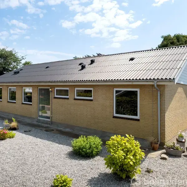 Et enkeltplans gult murstenshus med skråt tag. Huset har flere vinduer og en glasdør. Grusbelagt forhave med spredte grønne buske. Klar blå himmel i baggrunden.
