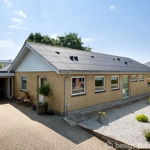 Et gult murstenshus med bølgeeternittag har en flisebelagt indkørsel og en grusbelagt have med planter. Et skilt står i haven med teksten "Til salg". Skraldespande ses nær huset.