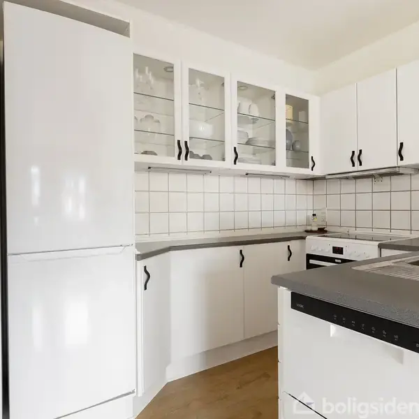 Et hvidt køkken med grå bordplade, skabsdøre med sorte håndtag, indbygget komfur og ovn. Væggene har hvide fliser. Der ses et montret billede i baggrunden.