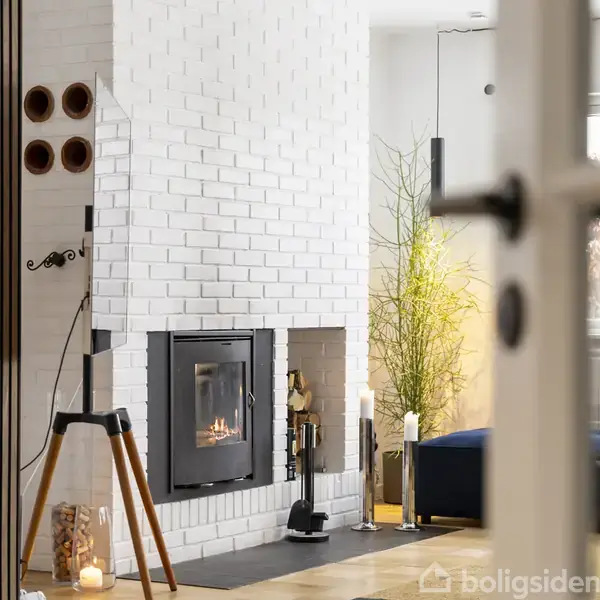 En brændeovn med flammer i en hvid murstensvæg står ved siden af dekorative genstande og en plante i en moderne stueindretning.