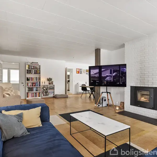 Stor, lys stue med blå sofa og moderne pejseindsats. Et tv står i rummet med åbne reoler og lyse vægge. Bøger og tidsskrifter fylder hylderne, og en lænestol står nært.