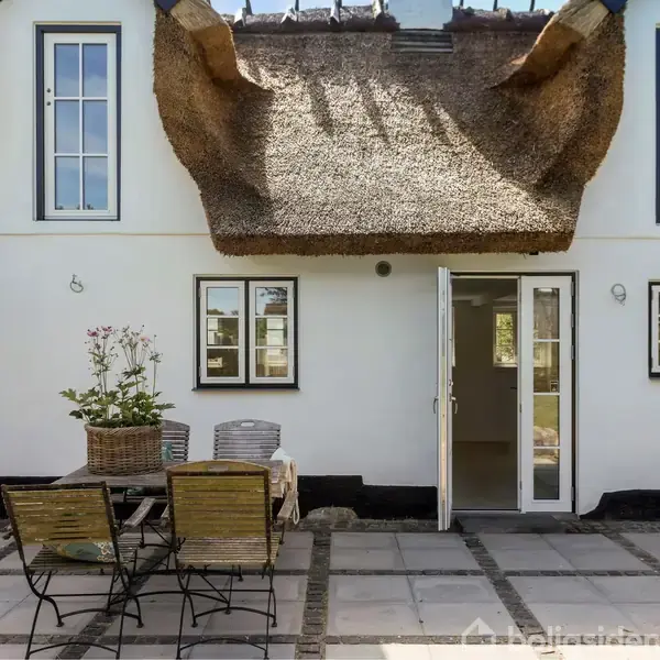 Et traditionelt hus med stråtag, åbne døre og vinduer. En terrasse med fliser rummer et bord med plantekurv og to stole. Sollys giver en lys atmosfære.