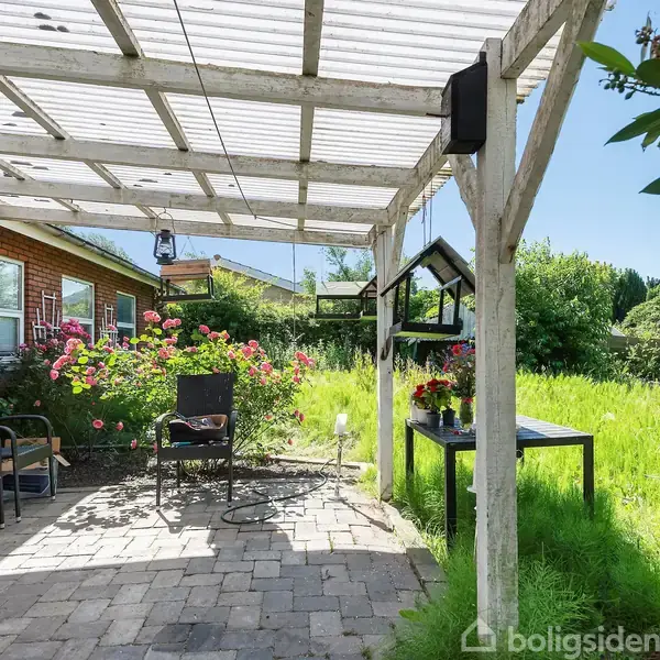En terrasse med hvide trælister som tag. To sorte stole står ved et bord. Blomster og grønne planter omgiver området med en murstensbygning i baggrunden.
