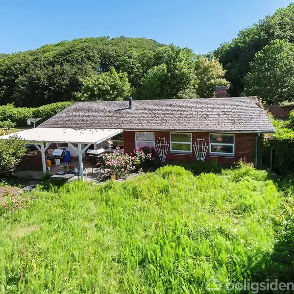 Et murstenshus står i en have med lang græs og en overdækket terrasse. Omkring huset er der frodig vegetation og baggrunden domineres af skovklædte bakker.