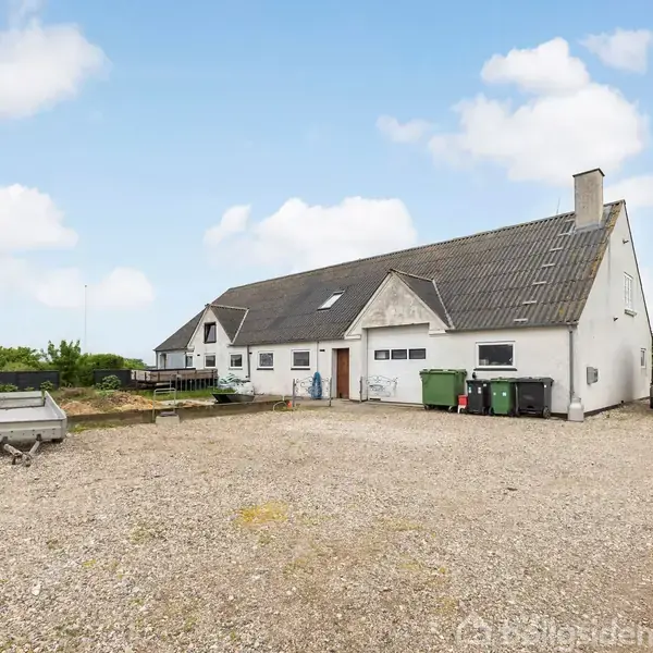 Hvidt hus med sort tag står på en grusbelagt gårdsplads med flere skraldespande og en trailer. Baggrunden viser en blå himmel med få skyer.