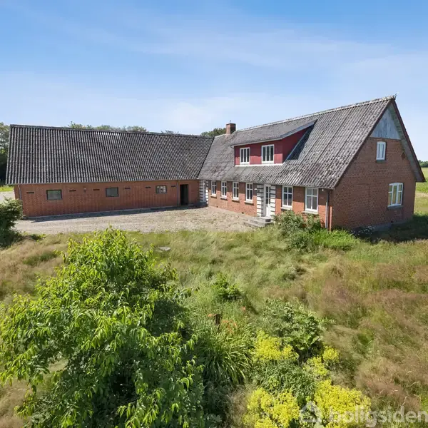 En stor, rød murstensgård står stille blandt frodige grønne marker. Det har et skråt tag og flere vinduer, der spejler den klare blå himmel.
