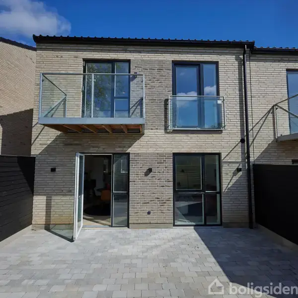 Rækkehus med to altaner og store vinduer, åbner op til en flisebelagt gårdhave. Sollys skinner på boligen, omgivet af murstensvægge og under en blå himmel.