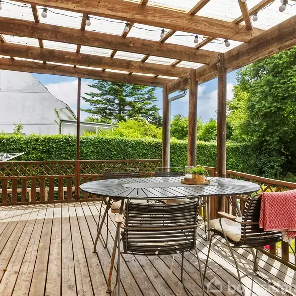 Rundt havebord med stole står på en træterrasse under en pergola. En pink plaid ligger på én stol. Haven er omkranset af buske og træer, mens en bagbygning er synlig i baggrunden.