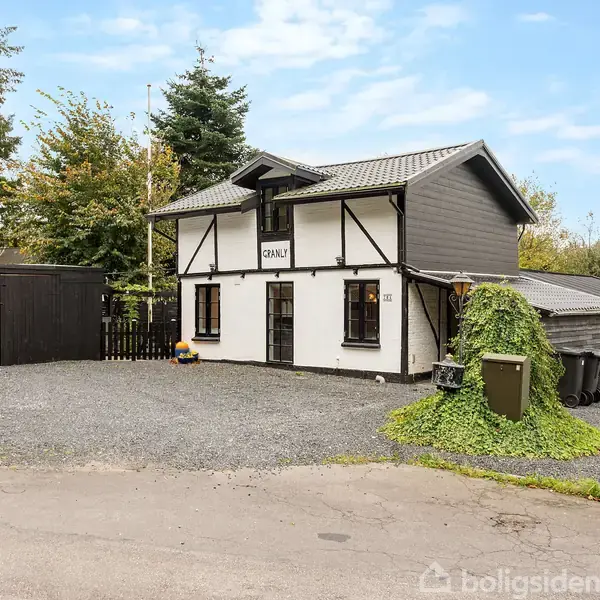 Et to-etagers hus står på en grusbelagt gårdsplads omgivet af træer. Skiltet på husets facade viser teksten "Chalet". En grå carport ligger ved siden af huset.