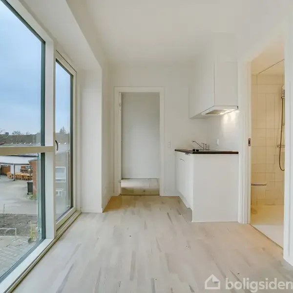 Et lyst, tomt rum med store vinduer til venstre og et lille hvidt køkken til højre. Udenfor ses en parkeringsplads med biler. En dør fører til et tilstødende rum.