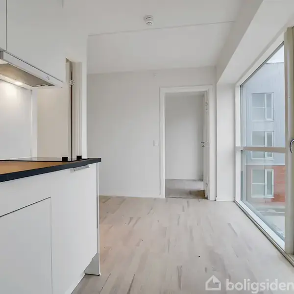 Et lyst, moderne køkken med hvidt inventar og sort bordplade står stille i et åbent rum med store vinduer, der giver udsigt til en bygning. Ingen tekst tilgængelig.