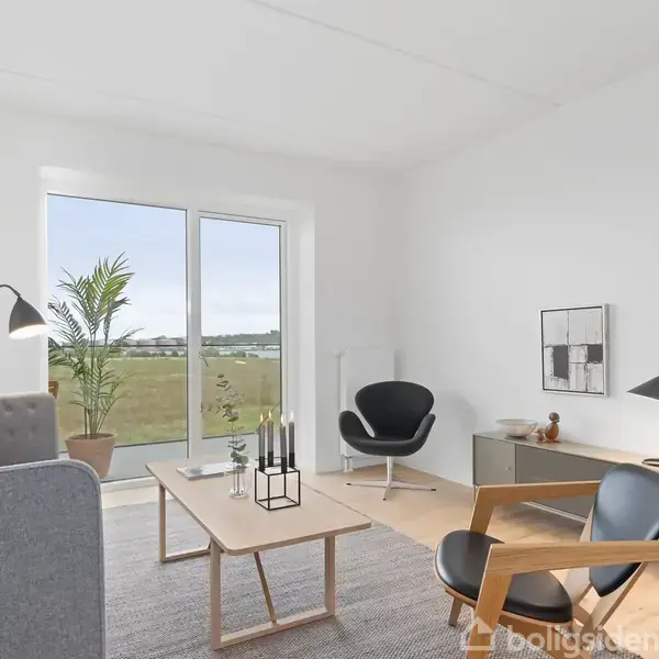 En lys stue indeholder en grå sofa, en træstol og et sofabord med lys dekor. En stor plante står ved terrassedøren, som åbner op til en udsigt over landskabet.