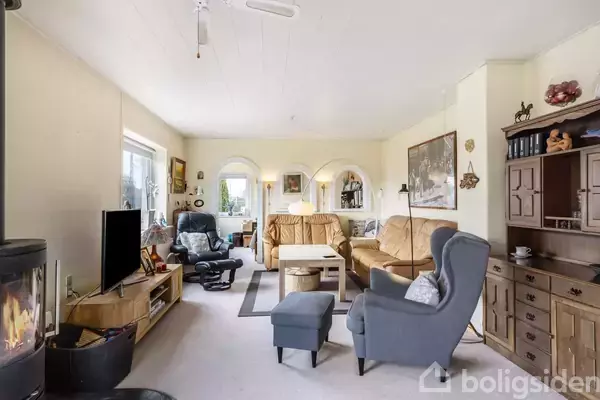 Stue med møbler, inklusiv sofaer, lænestole og sofabord, i hygge-kontinental stil. Brændeovn på venstre side og TV på et lavt bord. Store vinduer giver naturligt lys.