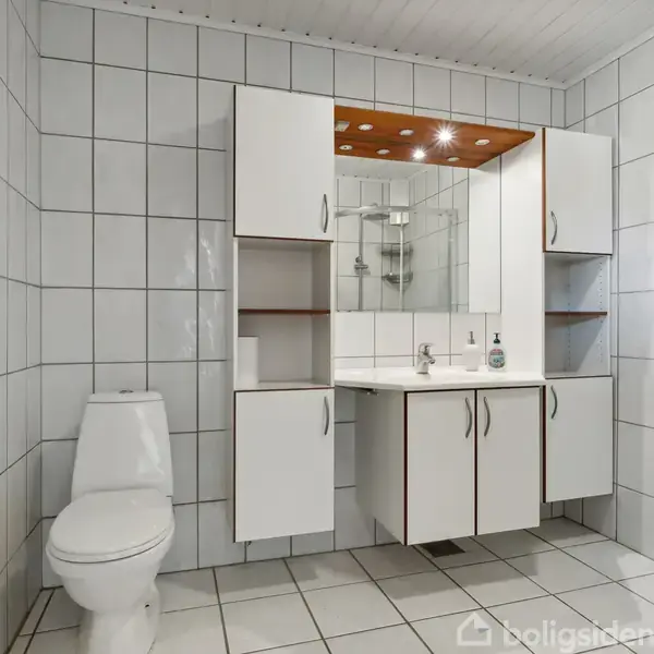 Et badeværelse indeholder en hvid toiletstol og hvidt bademøbel med spejl og skabe, omgivet af hvide fliser på vægge og gulv. En glasbruseniche er synlig i højre side.