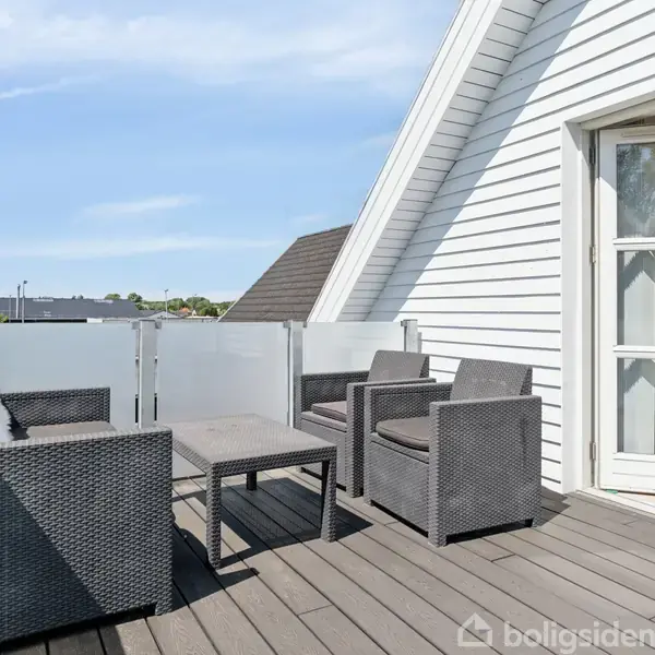 Terrassemøbler står på en moderne træterrasse. Møblerne inkluderer to stole og en sofa med et bord imellem. Terrassen er omgivet af glasværn og ved siden af en hvid bygning.