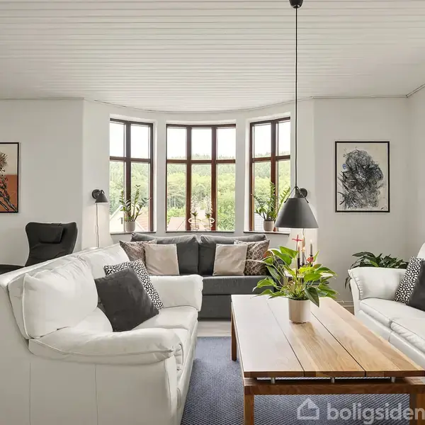 To hvide sofaer står omkring et træbord i en lys stue. Store vinduer i baggrunden slipper naturligt lys ind. Planter og moderne kunst dekorerer rummet.