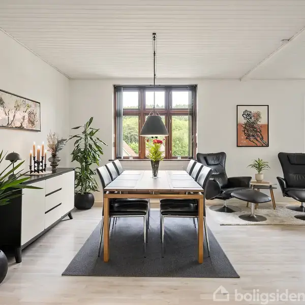 En spisestue med et træbord og otte stole er centralt placeret. På væggen hænger kunstværker, og store vinduer giver udsigt til grøn natur. Planter og stole fuldender rummet.
