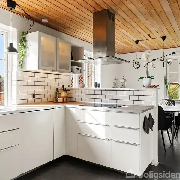 Køkken med hvide skabe og integreret komfur i hvidt flisebord, omgivet af lyse vægge og stort vindue. Træloftet giver en varm atmosfære. Spisebord med sorte stole ses i baggrunden.