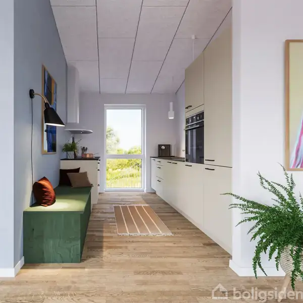 En smalt køkken med hvide skabe på højre væg, en grøn bænk med puder til venstre, en trælaminatgulv og et gulvtæppe. En glasdør viser en grøn udsigt udenfor.