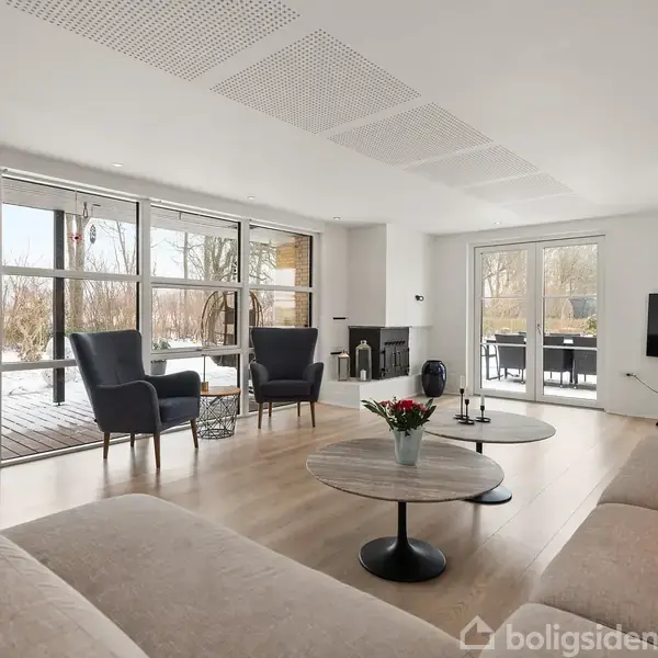 En stue med moderne møbler inkluderer en stor sofa, to lænestole, og et sofabord med blomster. Rummet har store vinduer, som giver udsigt til en træterrasse og have.