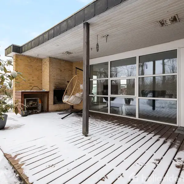 En overdækket terrasse i snevejr, hvor en gyngestol hænger ved siden af en åben pejs. Trægulvet er delvist dækket af sne.