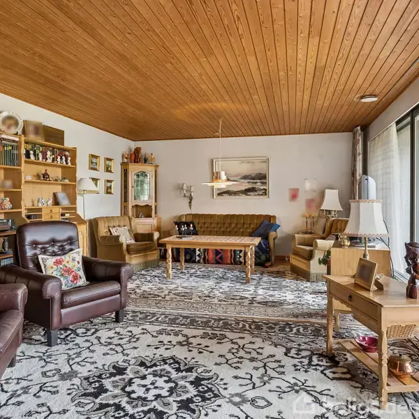 Stuen har brune læderlænestole og en beige sofa på en dekorativt mønstret tæppe. Træloftet og bøger i hylder skaber en hyggelig, klassisk atmosfære. Store vinduer giver lysindfald.