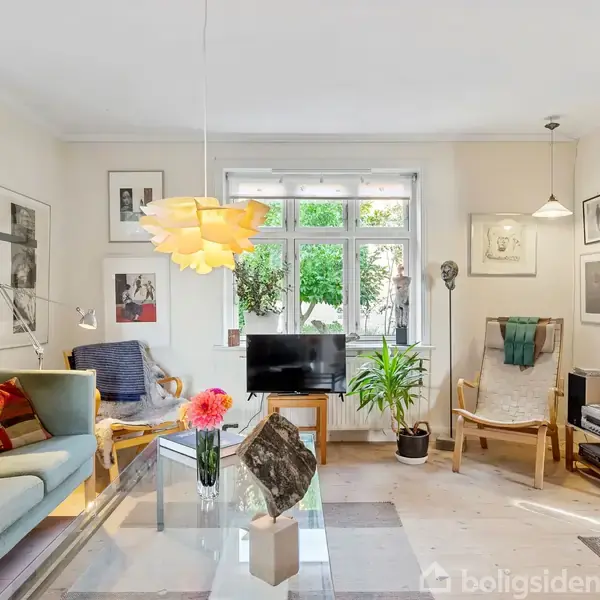 Stue med sofa og stole omkring sofabord, dekoreret med blomster. Kunst hænger på væggene. Et lille tv står foran vindue med udsigt til grønt område.