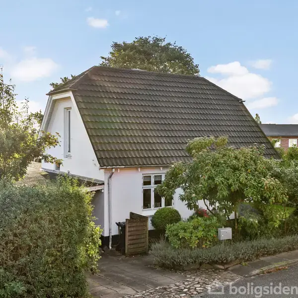 Huset, en hvid murstensbygning med brunt tag, står stille omgivet af frodig vegetation i et roligt nabolag med blå himmel og enkelte skyer.