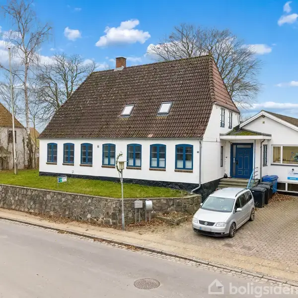 En hvid bygning med blå vinduer står ved en stenmur. En hvid bil er parkeret i indkørslen. Omgivet af andre huse og træer under en blå himmel.