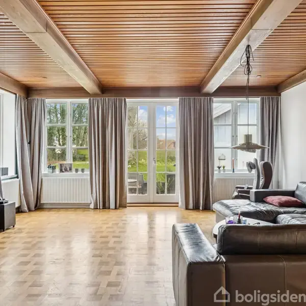 En stue med sort lædersofa står foran store vinduer og franske døre, der fører ud til en terrasse. Træloft og parketgulv skaber en hyggelig atmosfære.