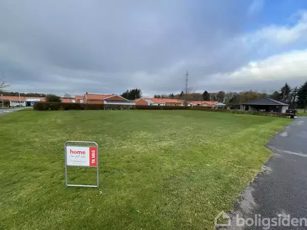 Et græsareal med et skilt, der viser "home TIL SALG" står på en plæne ved siden af en vej. Baggrunden har røde hustage og træer under en overskyet himmel.