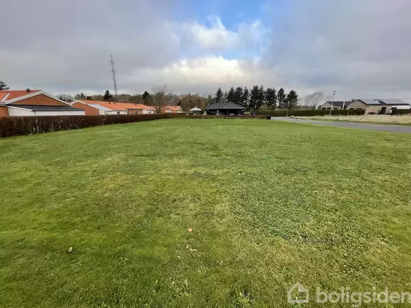Græsplæne strækker sig foran række af røde tage i et boligområde. Bagved er der træer og en overskyet himmel. Til højre ses en vej.