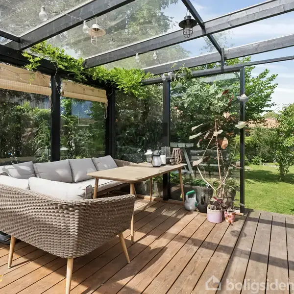 En komfortabel sofa står på en træterrasse under en glastag, omgivet af grønne planter og med et bord foran. Terrassen har udsigt til en frodig have.