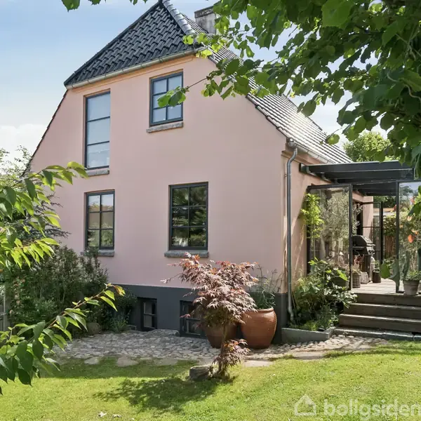 Et lyserødt hus med sort tegltag, omgivet af grønne planter og terrasse. Store vinduer ses på facaden, og en stenterrasse med potteplanter skaber en hyggelig indgang.