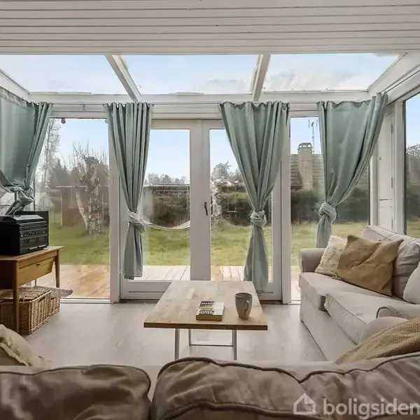 En lys stue med bløde sofaer vender mod en glasvæg med dobbeltdøre. Grønne gardiner og et bord med en kop ses forrest. Udsigt til have udenfor.