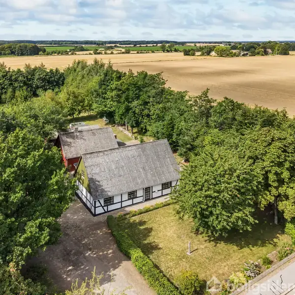 Bindingsværkshus med stråtag ligger i en frodig have, omgivet af træer. Baggrundens vidtstrakte marker og blå himmel skaber en rolig, landlig atmosfære.