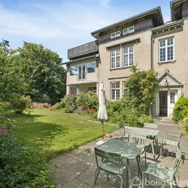En have med et havebord og stole står på en flisebelagt terrasse. Baggrunden er et stort hus med mange vinduer, omgivet af grønne buske og træer.