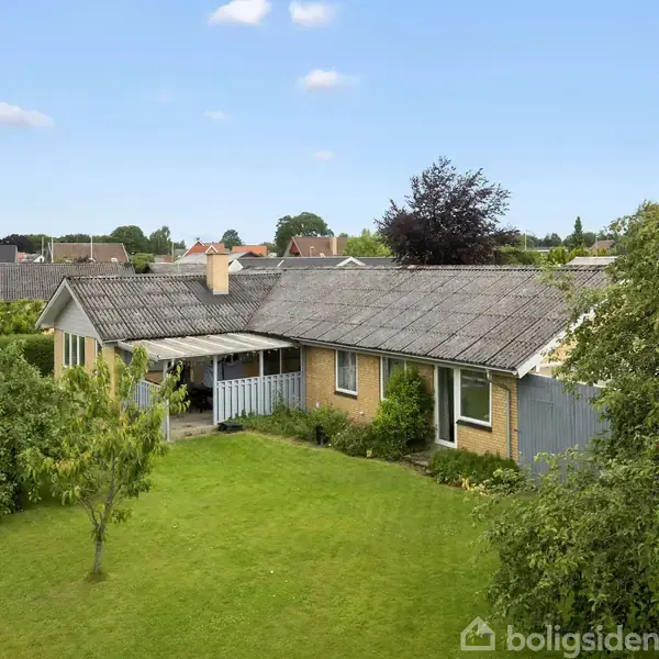 En gul murstensvilla med skrånende tag står i en frodig have med flere træer og en lille udestue. I baggrunden ses nabohuse og blå himmel.