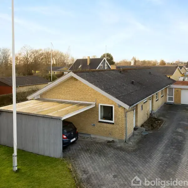 En gulmurstensvilla med en carport, hvor en bil holder parkeret. Villaen ligger i et boligområde omgivet af andre huse og grønne områder under en klar blå himmel.