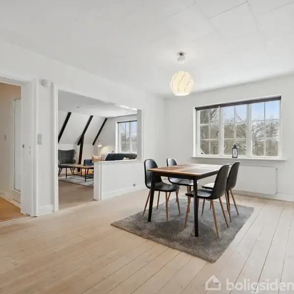 Spisebord med fire stole står på tæppe i lys stue. Et stort vindue bagved giver udsigt til træer. Til venstre ses en åbning mod en stue med sofa.