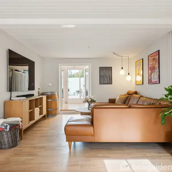 Brunt lædersofaarrangement står i en stue med trælaminatgulv og vægkonsoller. Et stort TV hænger på væggen, og rummet er dekoreret med planter, kunst på væggen og en stor døråbning mod en lysere udendørs terrasse.