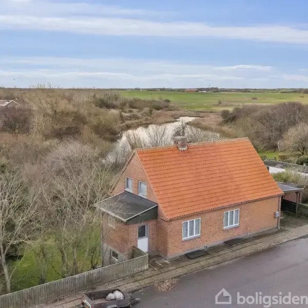 Murstenshus med rødt tegltag står stille langs en vej, omgivet af træer og buske, i et landligt område med åbne marker i baggrunden.